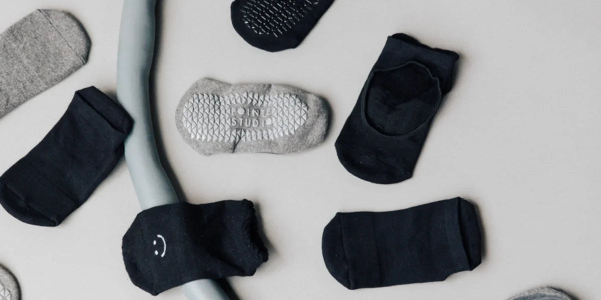 Billedet viser forskellige yoga- og pilatessokker fra Pointe Studio. Stilarter som ses inkluderer grå sokker med hvide greb, sorte sokker med smilende ansigt og andre forskellige designs. Disse sokker er designet til at give greb og komfort under yoga og pilates.